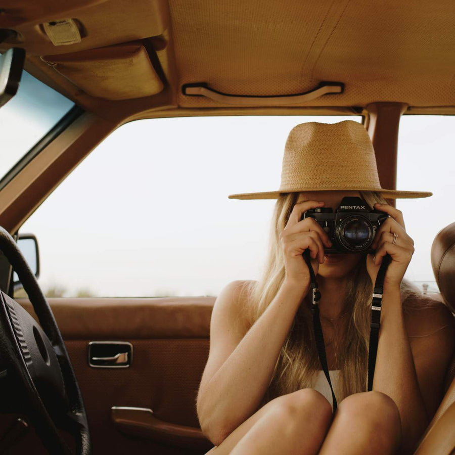 woman in a wide brim straw hat taking a photo with a film camera from a car